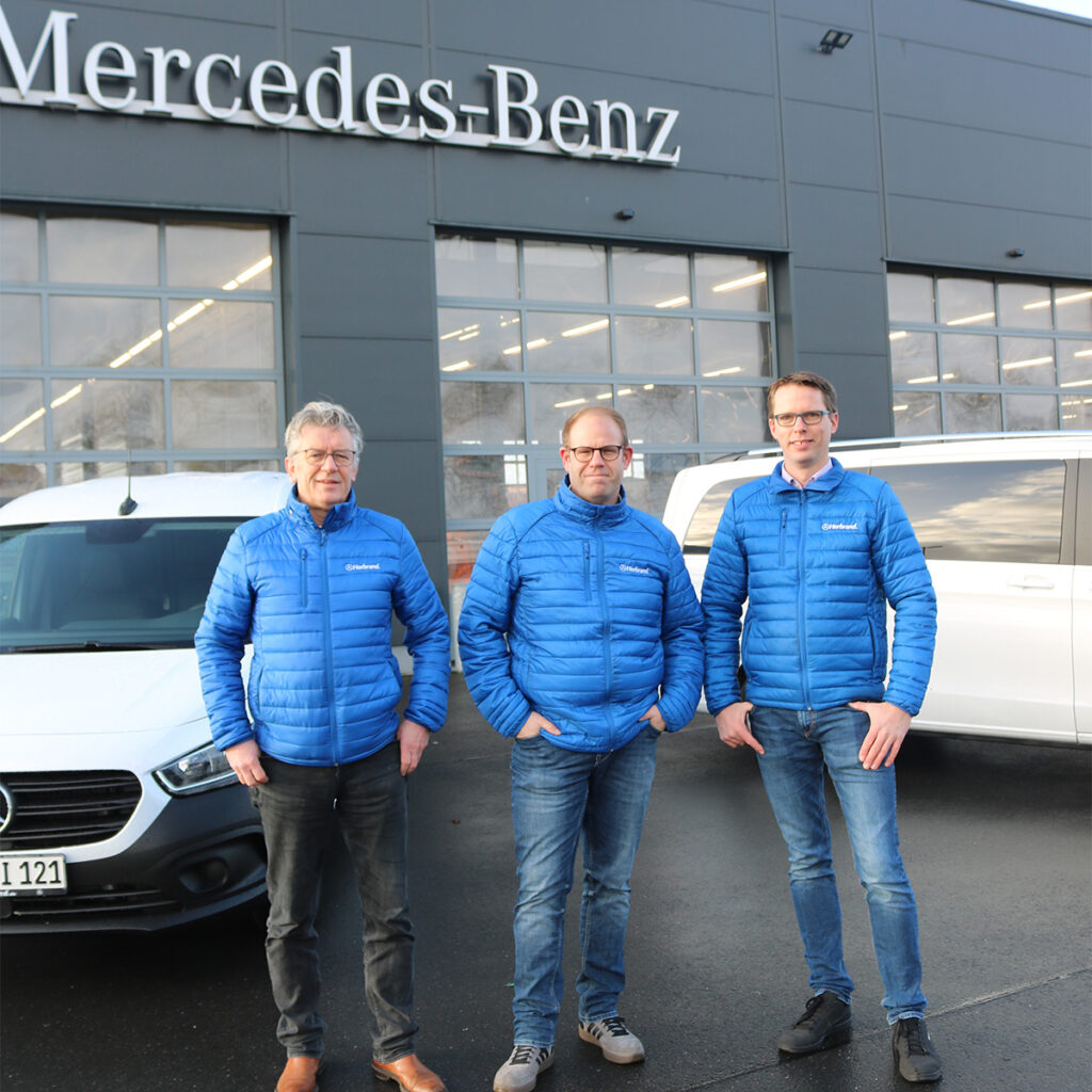 Drei Männer in blauen Herbrand-Jacken stehen vor einem Mercedes-Benz Autohaus, lächelnd in die Kamera.