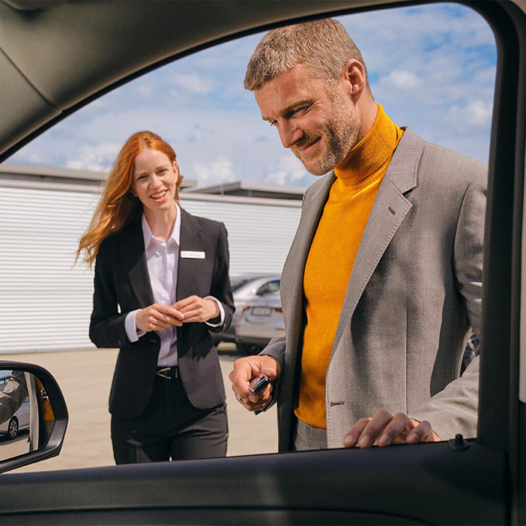 Ein lächelnder Mann in einem grauen Anzug und orangefarbenem Rollkragenpullover nimmt Autoschlüssel von einer freundlichen, rotblonden Verkäuferin entgegen. Die Szene spielt sich vor einem Autohaus ab, im Hintergrund sind geparkte Autos sichtbar. Beide Personen stehen außerhalb eines Fahrzeugs, und die Verkäuferin trägt eine dunkle Business-Jacke mit Namensschild.