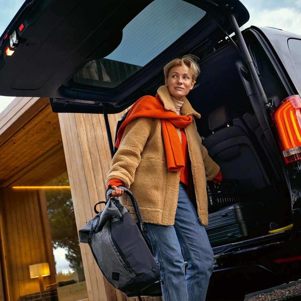 Eine Frau lädt Gepäck in einen Mercedes-Benz Transporter vor einem modernen Gebäude ein, Symbol für flexible und komfortable Transportlösungen.