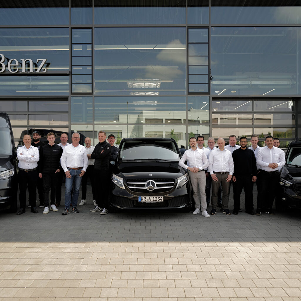 Eine Gruppe von Mitarbeitern in weißen Hemden steht vor einem Mercedes-Benz Autohaus, flankiert von schwarzen Mercedes-Benz Fahrzeugen.