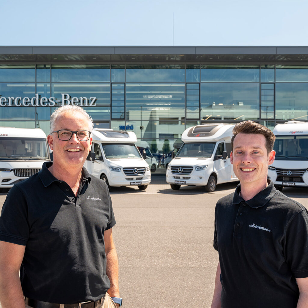 Zwei Verkaufsberater stehen vor dem Mercedes-Benz Autohaus Herbrand, umgeben von einer Auswahl an Reisemobilen. Sie lächeln und posieren in einer freundlichen Atmosphäre, was Teamgeist und Professionalität vermittelt.