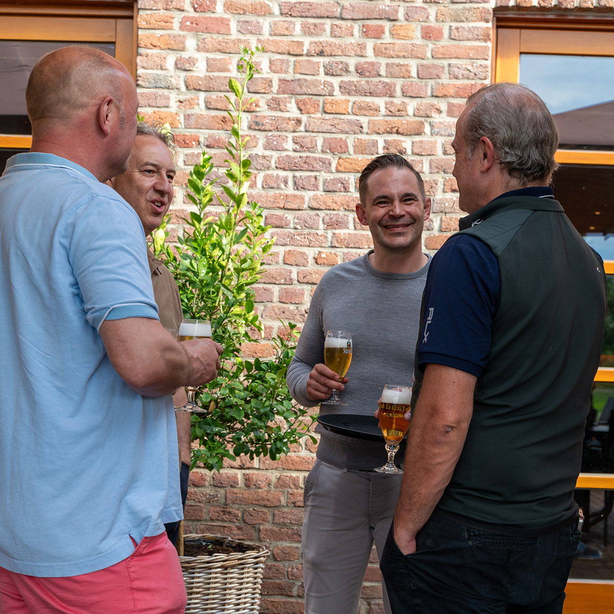 Vier Männer stehen draußen vor einem Backsteingebäude und unterhalten sich, jeder hält ein Glas Bier in der Hand.