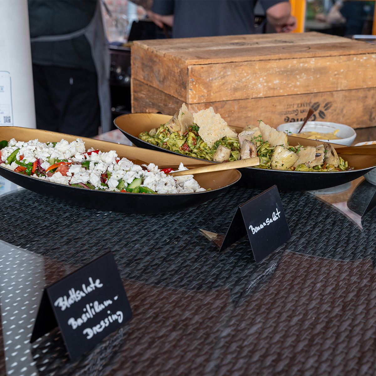 Zwei Salate auf einem Tisch in schwarzen Schalen, einer mit Feta und Gemüse, der andere mit Nudeln und Pilzen. Im Hintergrund ein Holzblock und weitere Personen.