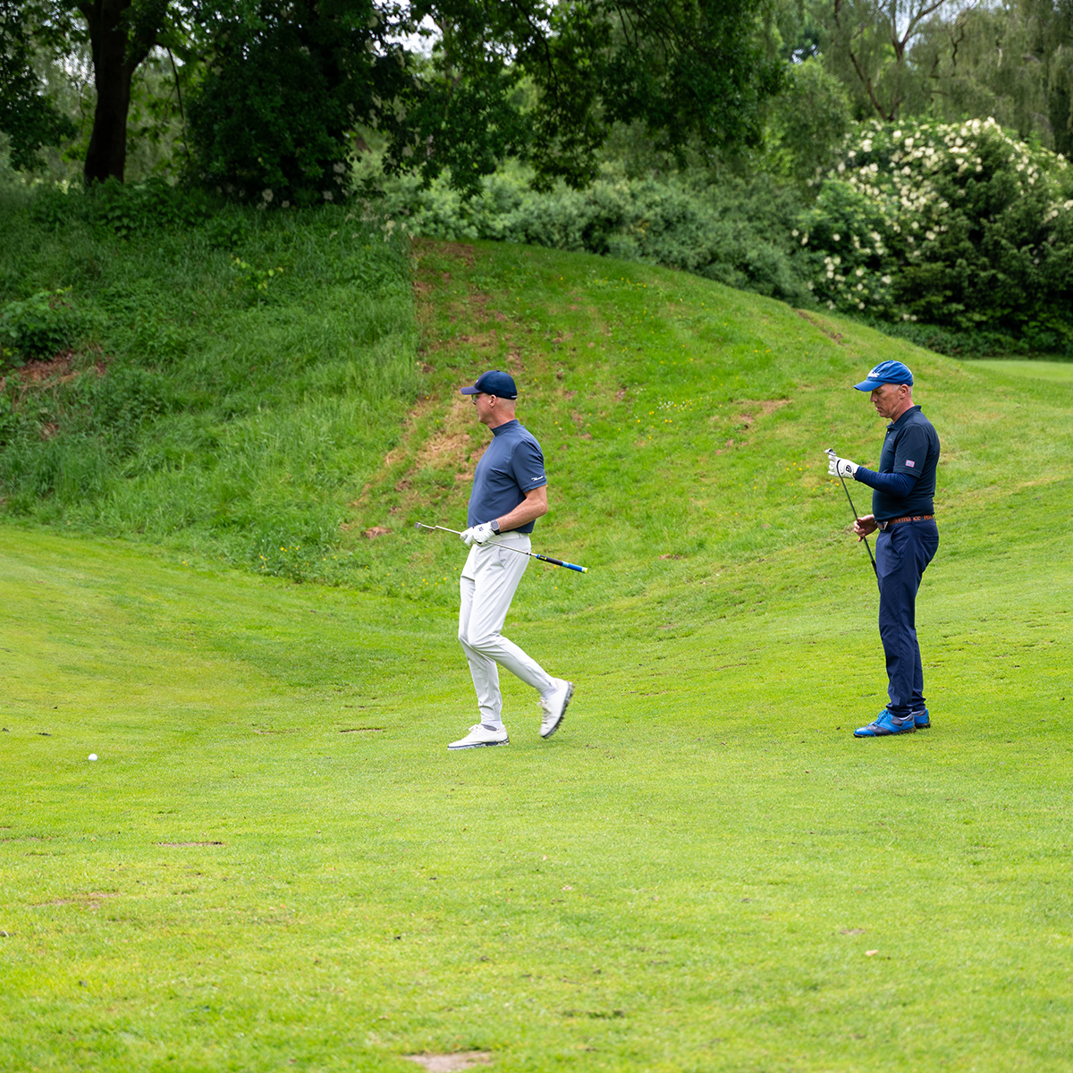 Zwei Golfer in sportlicher Kleidung, die auf einem Golfplatz nebeneinander stehen und ihre Schläger halten. Der Rasen ist grün und gepflegt.