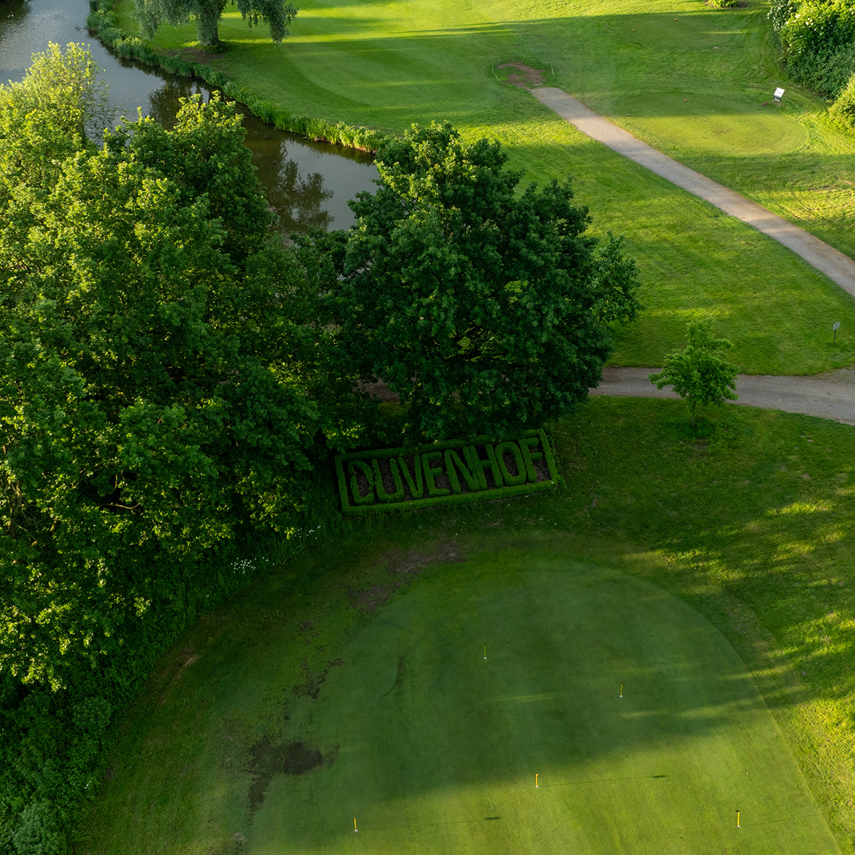 Luftaufnahme eines Golfplatzes mit gepflegtem Rasen, einem Weg und einem kleinen Teich. Im Vordergrund ist der Schriftzug "DUVENHOF" in die Hecke geschnitten.