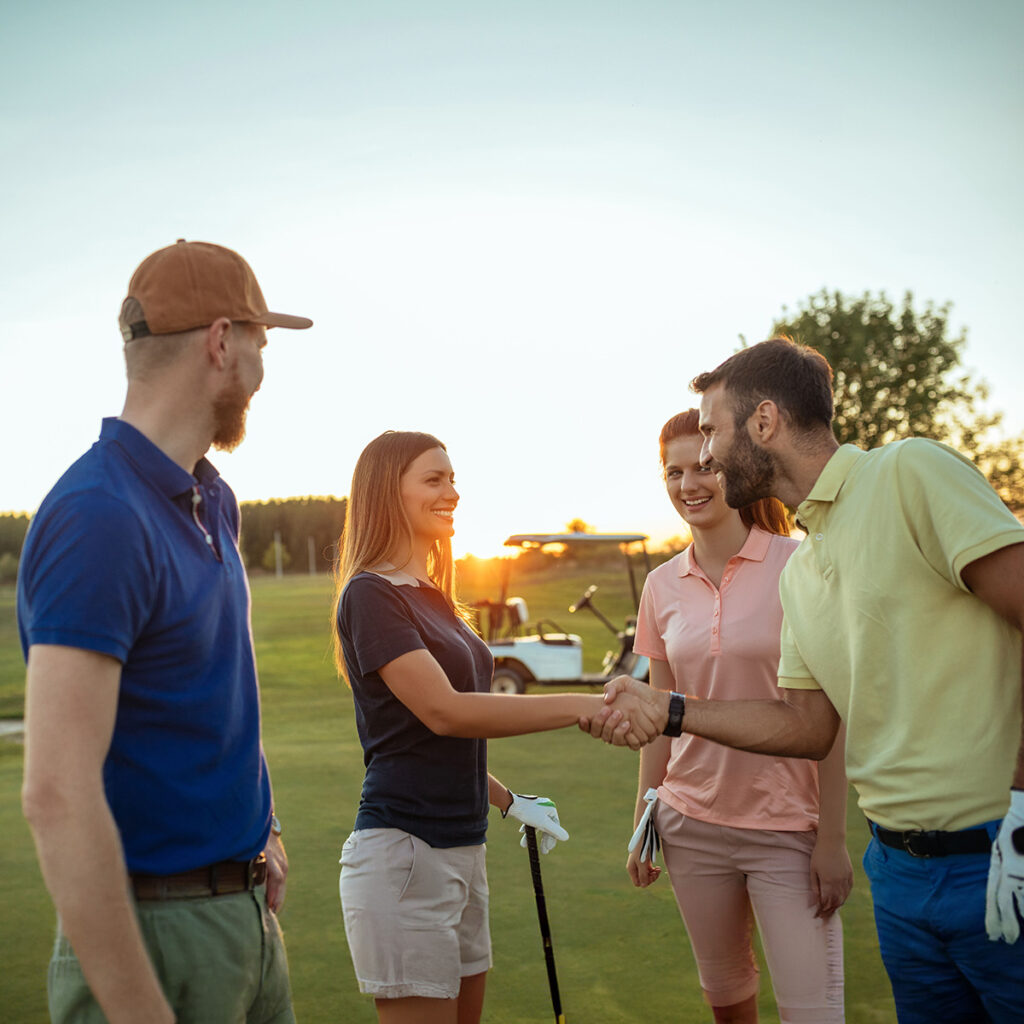Vier Golfer auf einem Platz bei Sonnenuntergang, die sich die Hand schütteln und lächeln. Ein Golfwagen steht im Hintergrund.