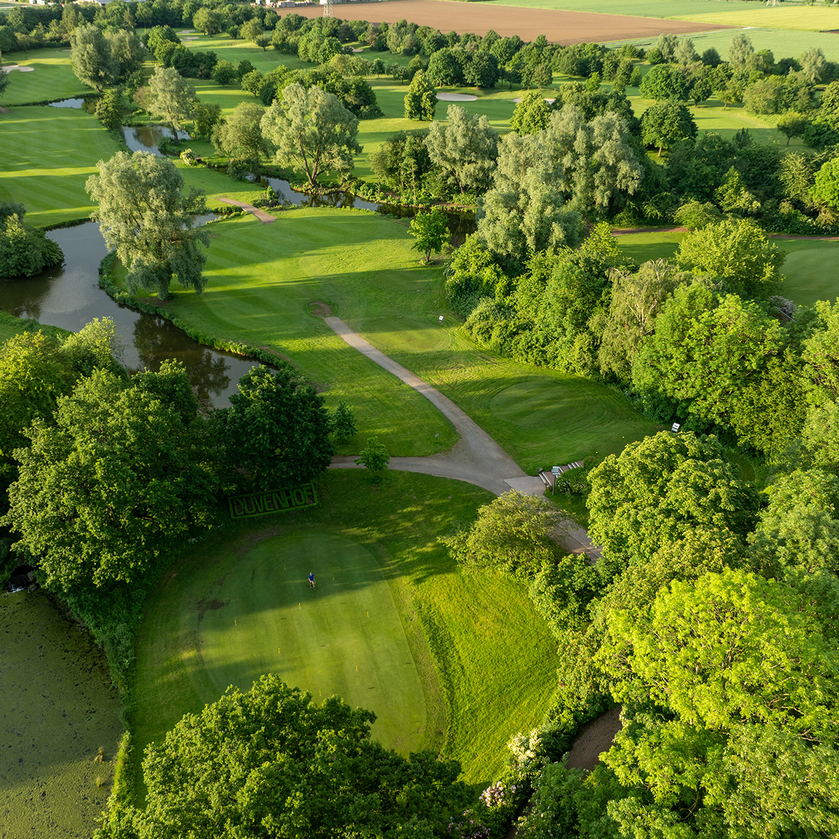 Luftaufnahme eines gepflegten Golfplatzes mit grünen Fairways und Bäumen im Hintergrund, die die malerische Landschaft umrahmen. Im Vordergrund ist ein Golfer auf dem Grün zu sehen.